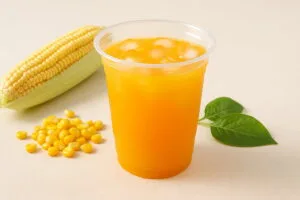 Compostable PLA clear cups with colorful drinks displayed beside corn, sugarcane, and PLA resin pellets on a beige background, symbolizing plant-based eco-friendly materials.