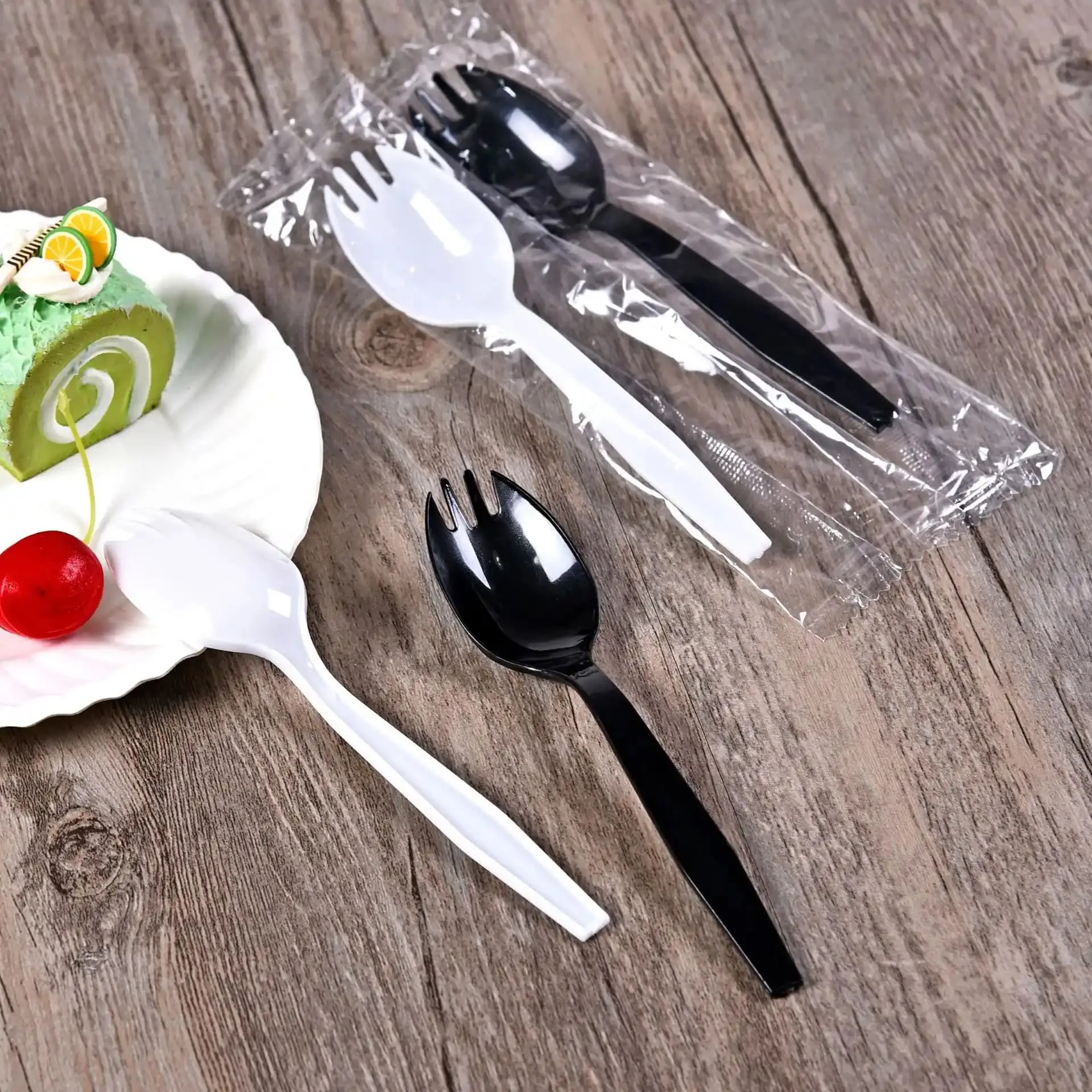 Cubiertos tradicionales de plástico PP y PS - Utensilios de un solo uso Cubiertos de plástico tradicionales fabricados con PP y PS, incluidos los cubiertos desechables en blanco y negro envueltos en plástico sobre una mesa de madera.