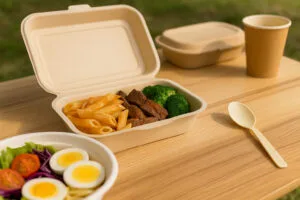 Compostable bagasse clamshell container with pasta and vegetables, cornstarch spoon, and paper cup on an outdoor picnic table.